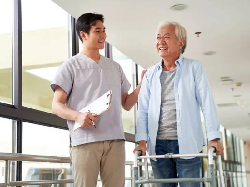 A therapist walking beside an older man steadying himself on a walker in a bright corridor