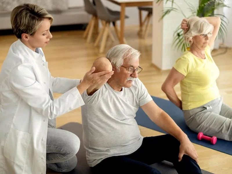 A therapist guiding an older man through a seated stretch on an exercise mat, with another patient exercising nearby