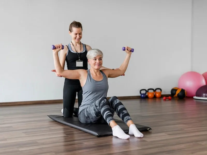 A therapist supporting a woman's arms as she lifts light dumbbells during a seated floor exercise