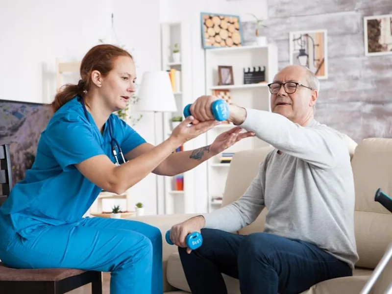 A therapist in blue scrubs supporting an older man's arm as he raises a light dumbbell overhead while seated